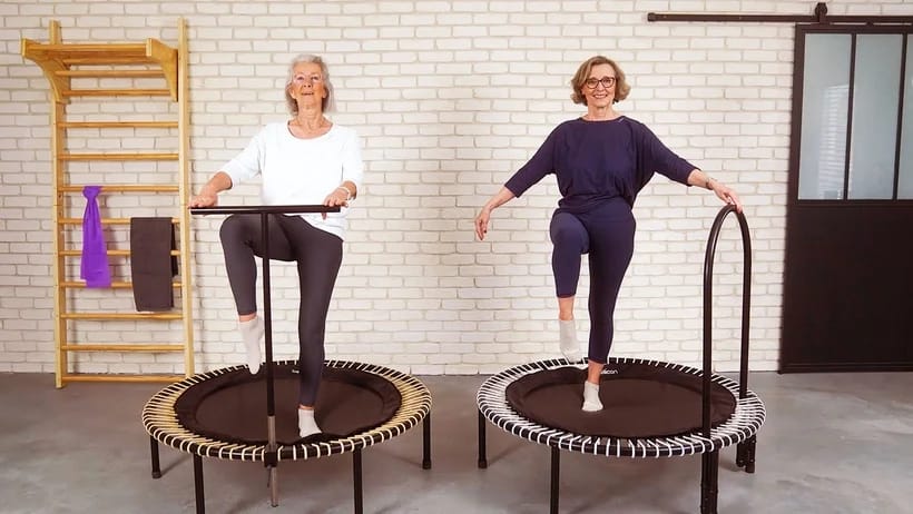 Zwei ältere Damen trainieren auf dem Minitrampolin, eine hält sich an der T-bar fest, die andere an der seitlichen Haltestange.